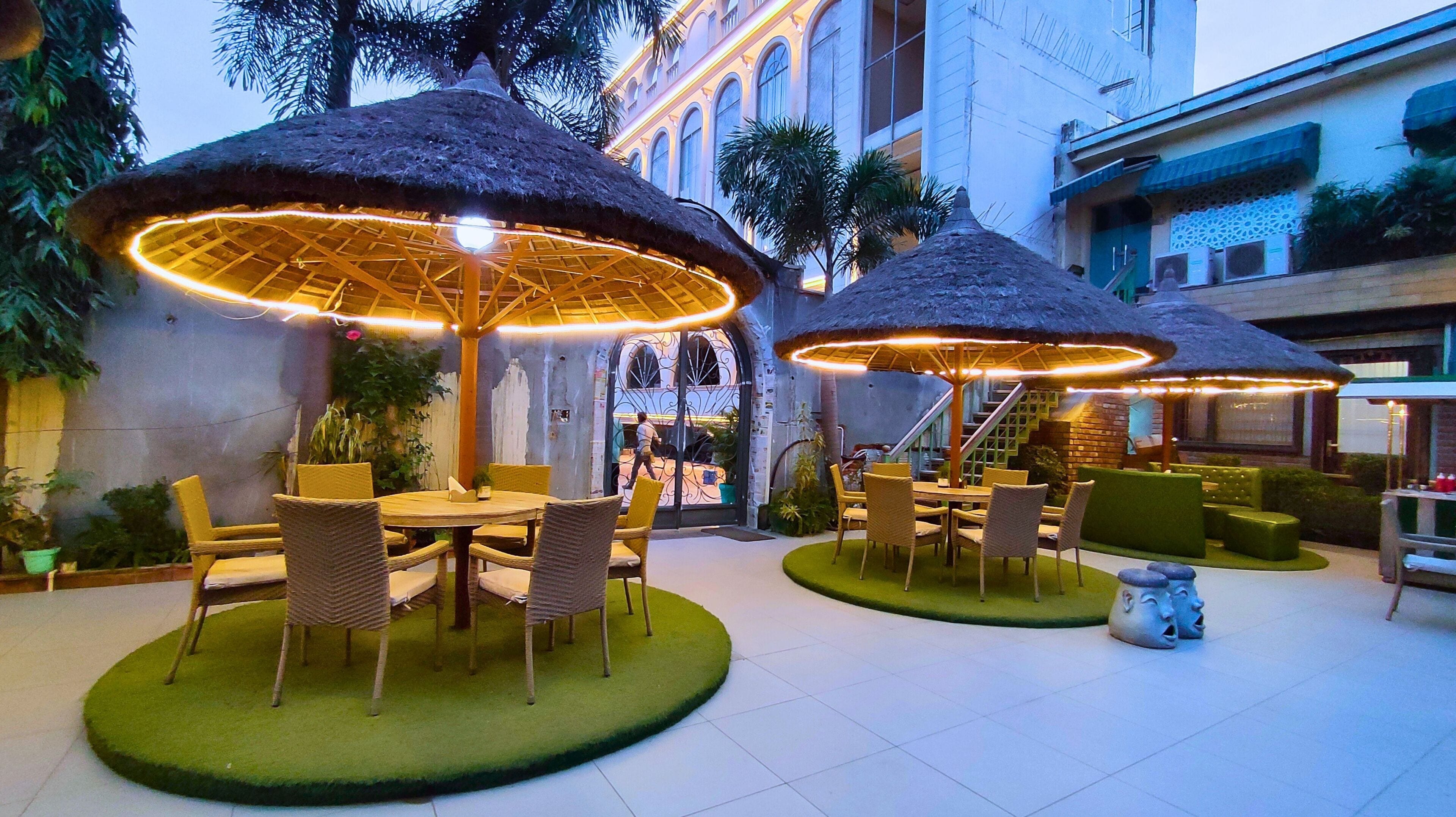 Un área de comedor al aire libre con mesas y sillas redondas, rodeada de césped artificial verde, y cubierta por sombrillas de paja.