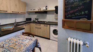 A compact kitchen with wooden cabinets, a microwave, a kettle, and a washing machine. A chalkboard with a welcome message is mounted on the wall. - The Arena - Apartment in the Center. Ideal for sightseeing. Metro 100m away (Napoli)