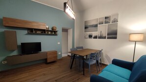 A modern living room with a wooden floor, a blue sofa, a dining table with chairs, a TV on a wooden stand, and wall-mounted shelves with decorative items. - The Arena - Apartment in the Center. Ideal for sightseeing. Metro 100m away (Napoli)