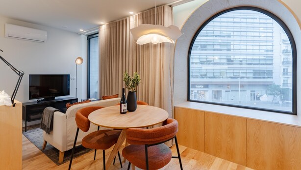 A modern living room with a round dining table, orange chairs, a TV, and a large window with a view of a cityscape. - Feel Porto Corporate Housing Boavista V (Porto)