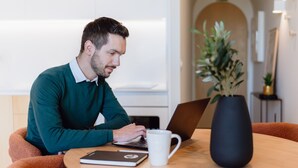 Une personne est assise à une table, devant un ordinateur portable, avec un carnet, une tasse et une plante en pot.