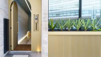 Um interior moderno com um corredor que leva a uma porta, um painel de plantas fixado na parede e uma grande janela com persianas.