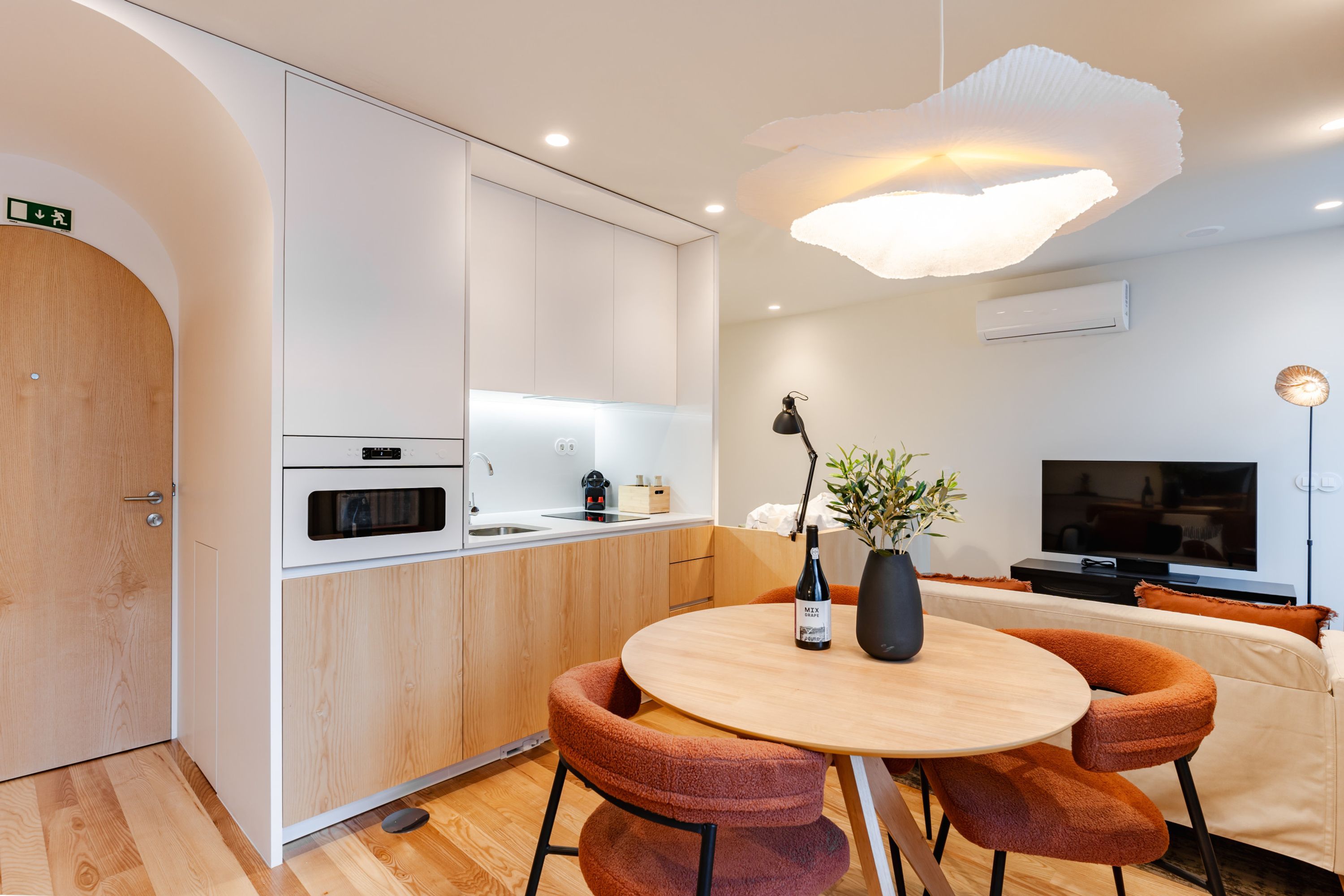 A modern kitchen with white cabinets, a microwave, and a sink. There's a dining area with a round table and orange chairs. A bottle of wine and a vase with a plant are on the table.