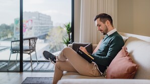 A person sitting on a couch with a laptop, a window with a view of buildings, and a plant on the windowsill.