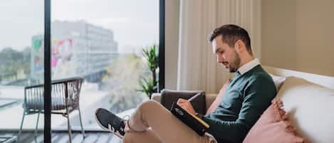 Une personne assise sur un canapé, un ordinateur portable sur les genoux, une fenêtre donnant sur des immeubles et une plante sur le rebord de la fenêtre.