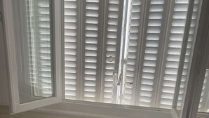 Ein Zimmer mit Fenster, an dem horizontale Jalousien angebracht sind, zwei Metallstühle und eine weiße Wand mit Lüftungsgitter.