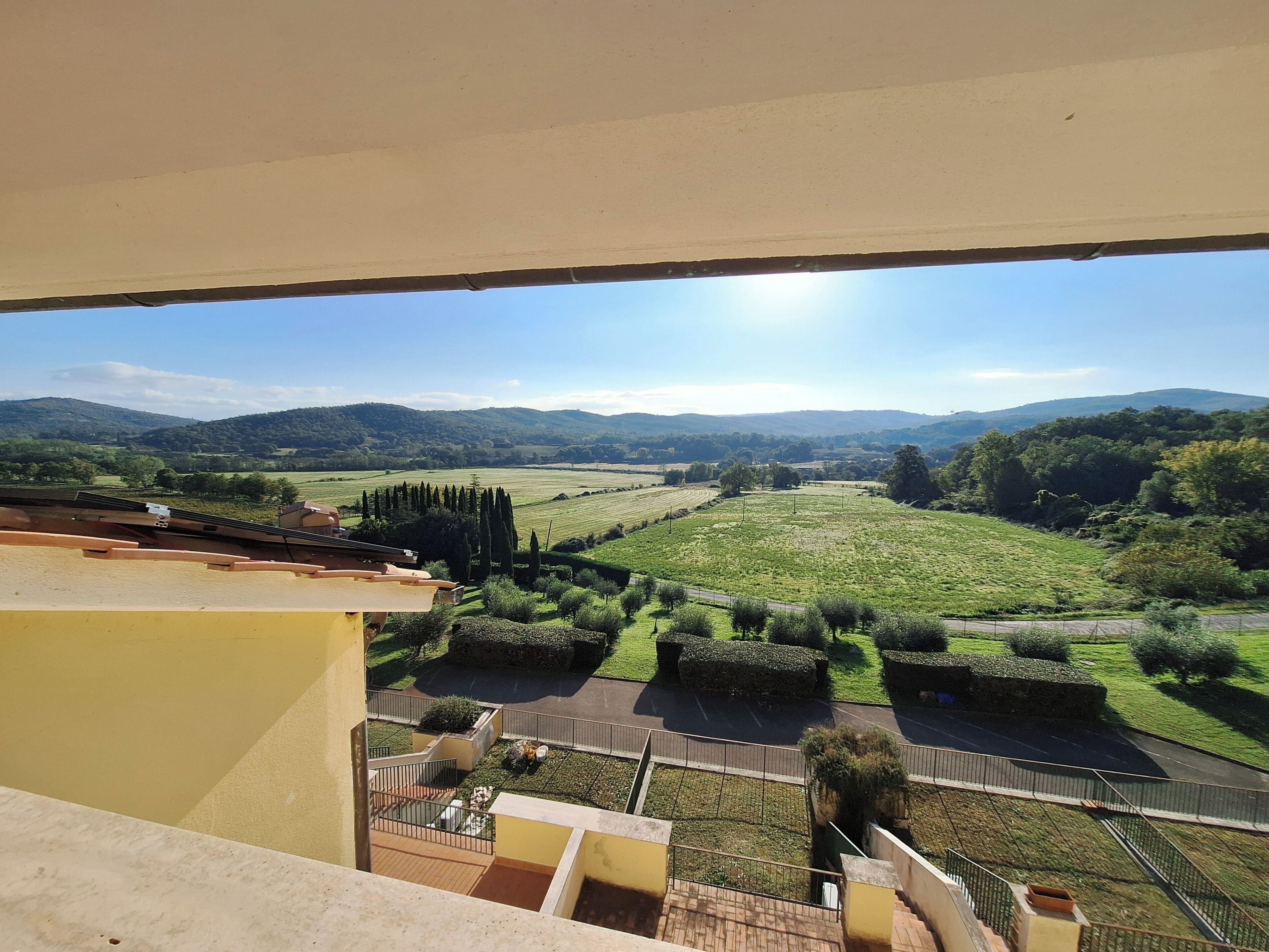 A view from a balcony overlooking a green landscape with trees and a road.