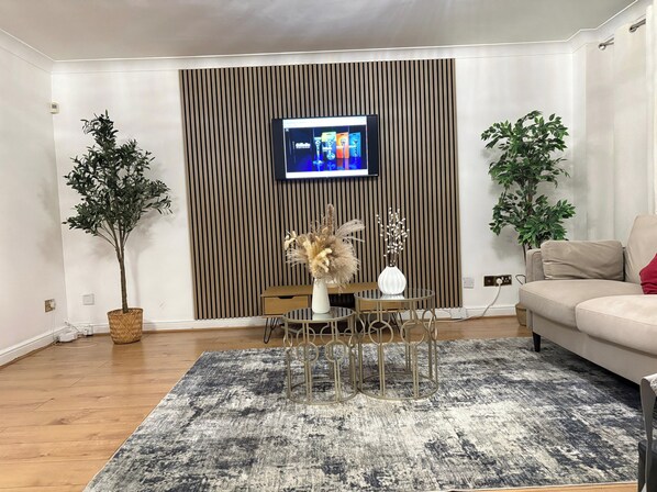 Ein Wohnzimmer mit Fernseher an einer Wand aus Holzpaneelen, einem runden Couchtisch mit einer Vase, einer Couch und zwei Blumentöpfen.
