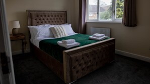 A neatly made bed with a green bedspread and white linens, a bedside lamp, and a view of the outside through a window with curtains. - Sleeps8-kingbeds-sofabed-garden-aircon-freeparking (London)