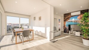A modern living room with a dining area, a flat-screen TV, and a large window offering a city view. - GuestReady - Peaceful stay in Charneca da Caparica (Charneca de Caparica)