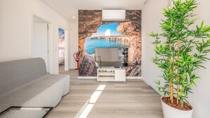 A modern living room with a grey sofa, a flat-screen TV on a white stand, a large wall mural of a coastal scene, and a bamboo plant in a white pot. - GuestReady - Peaceful stay in Charneca da Caparica (Charneca de Caparica)