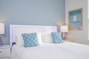 Una cama bien hecha con ropa blanca y dos almohadas decorativas azules, una mesita de noche blanca con una lámpara, y una obra de arte enmarcada en la pared.