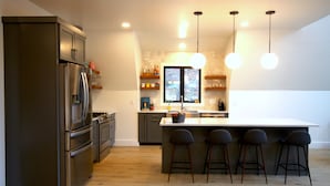 Une cuisine moderne dotée d’un îlot central, de tabourets de bar, d’appareils électroménagers en acier inoxydable et de luminaires suspendus.