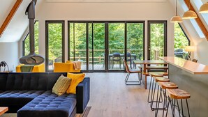 A modern loft-style living area with a large window, a black sofa, wooden bar stools, and a dining table. - Cozy A-Frame Cabin - In-Deck Hot Tub - near Boone (Vilas)