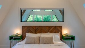 A bedroom with a large bed, wooden headboard, two bedside tables, and a window with a view of trees. - Cozy A-Frame Cabin - In-Deck Hot Tub - near Boone (Vilas)