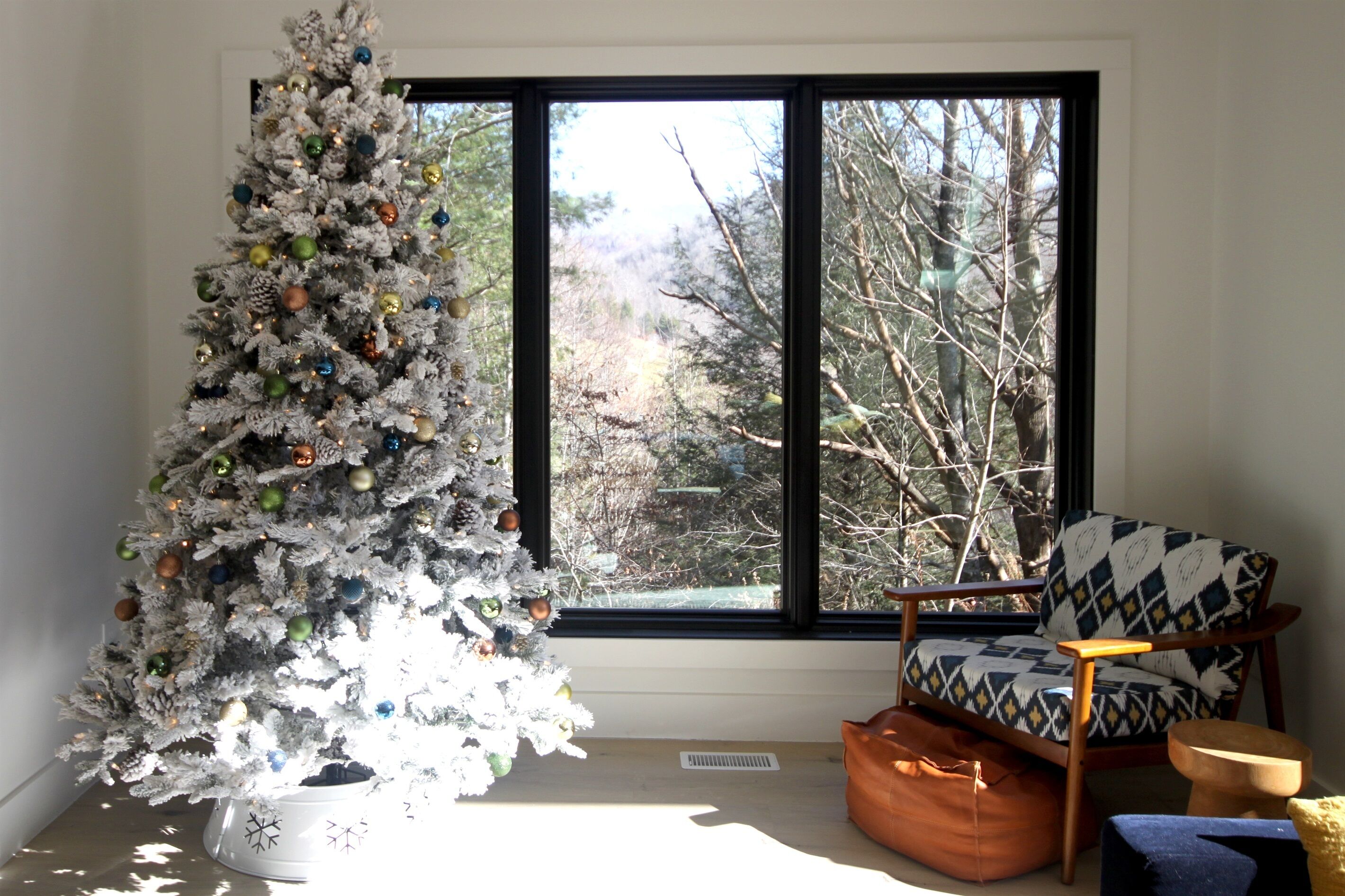 Een witte kerstboom met decoraties, een raam met uitzicht op bomen, een gestreept fauteuil en een houten bank.