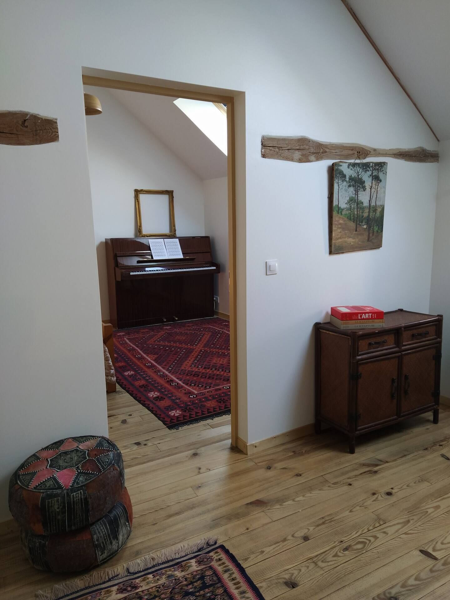A room with a wooden floor, a red patterned rug, a small round ottoman, a dark wooden cabinet, and a painting on the wall.