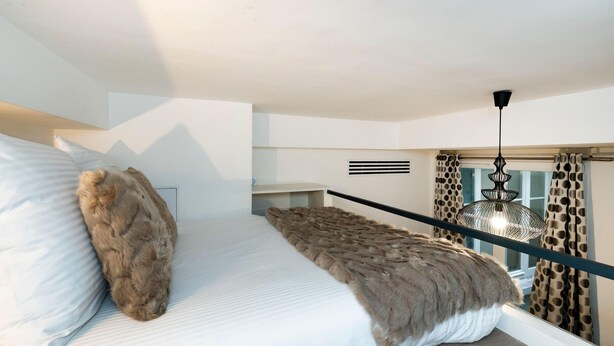 A bedroom with a bed, a window with curtains, a hanging light fixture, and a small shelf.