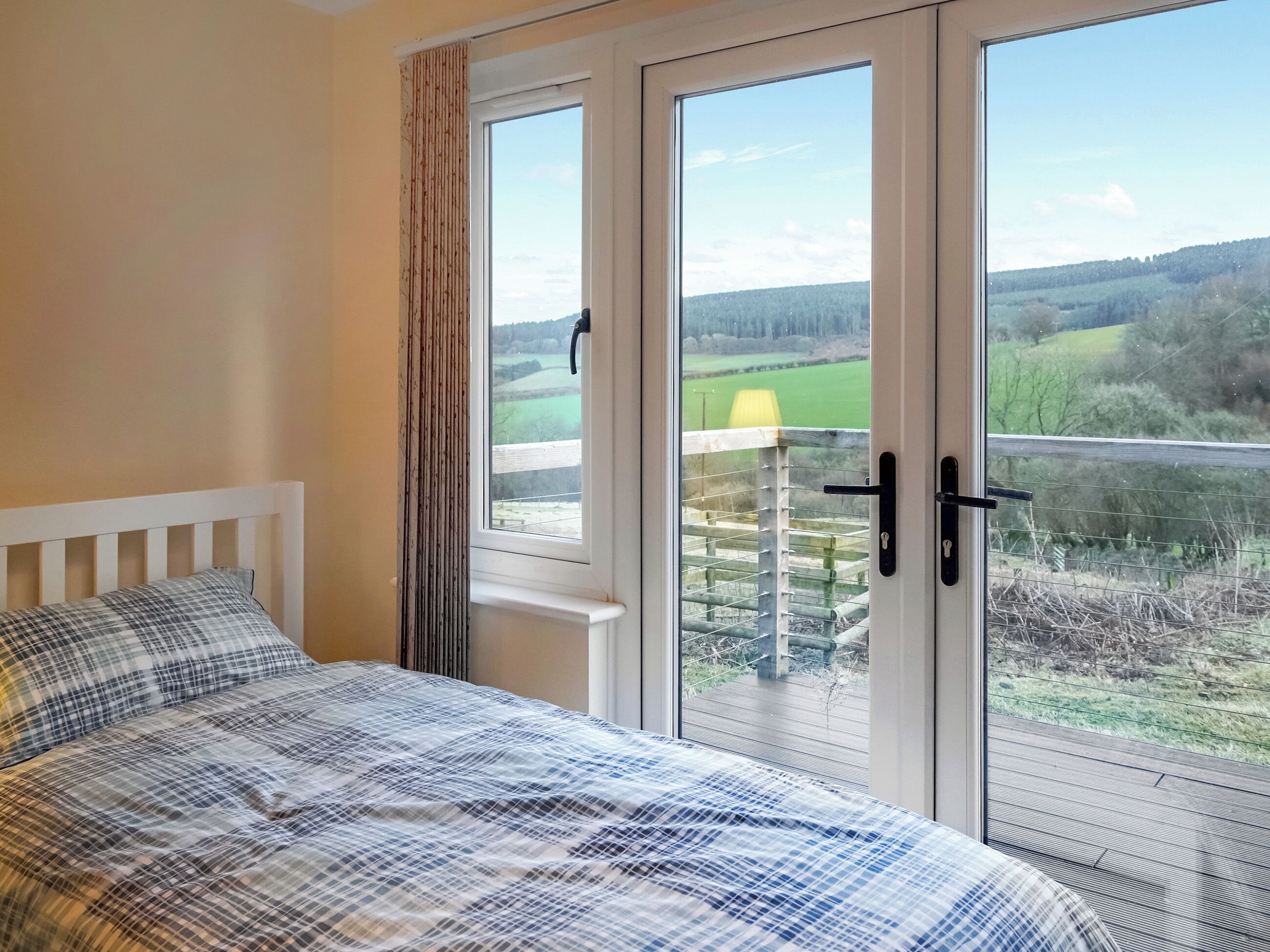 A bedroom with a bed, a window with a view of a green landscape, and a balcony with a railing.
