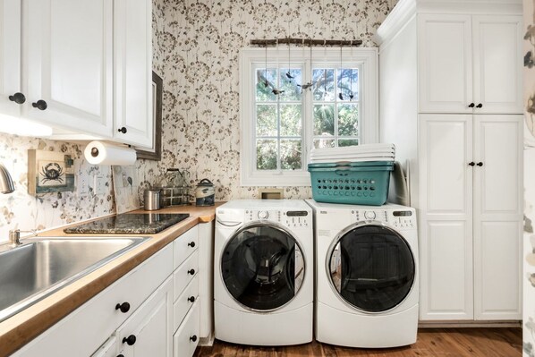 A laundry room with two front-loading washing machines, a window with curtains, and a wooden countertop.