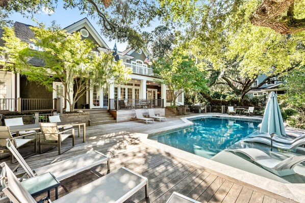 A pool area with lounge chairs and a hot tub, surrounded by trees and a house with a balcony. - 130 Flyway Drive - Villa ESTE - Pool heat optional (Kiawah Island)