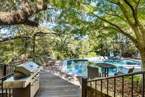 A wooden deck with a barbecue grill, a swimming pool with lounge chairs, and a view of the surrounding trees. - 130 Flyway Drive - Villa ESTE - Pool heat optional (Kiawah Island)