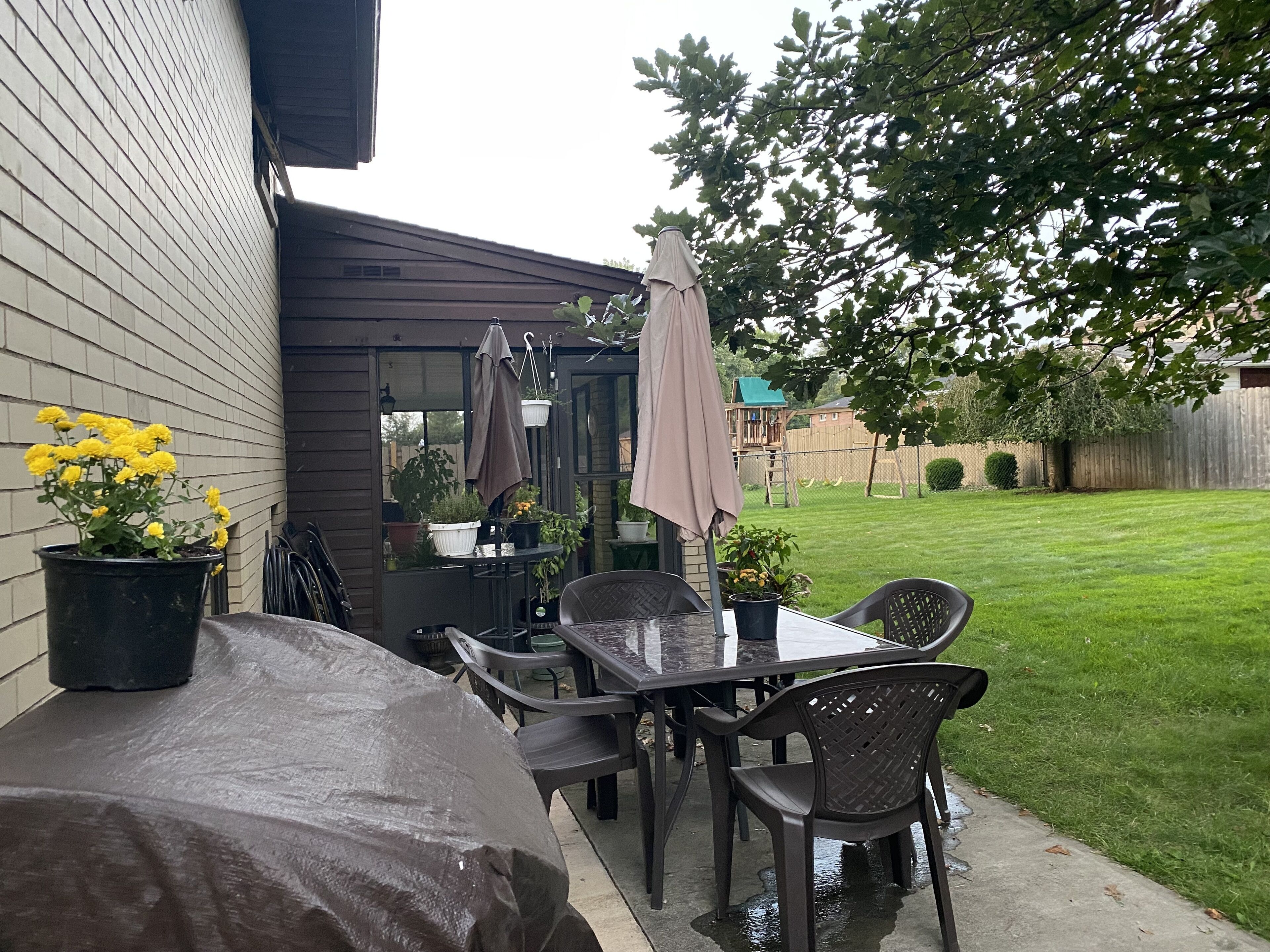 An outdoor patio with a covered table and chairs, surrounded by potted plants and a view of a grassy yard.