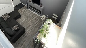 A modern living room with a grey wall, a flat-screen TV mounted above a fireplace, a black sofa with patterned cushions, and a dining area with a table and chairs. - The Cottages@Gull&Castle#4 (Wasagaming)