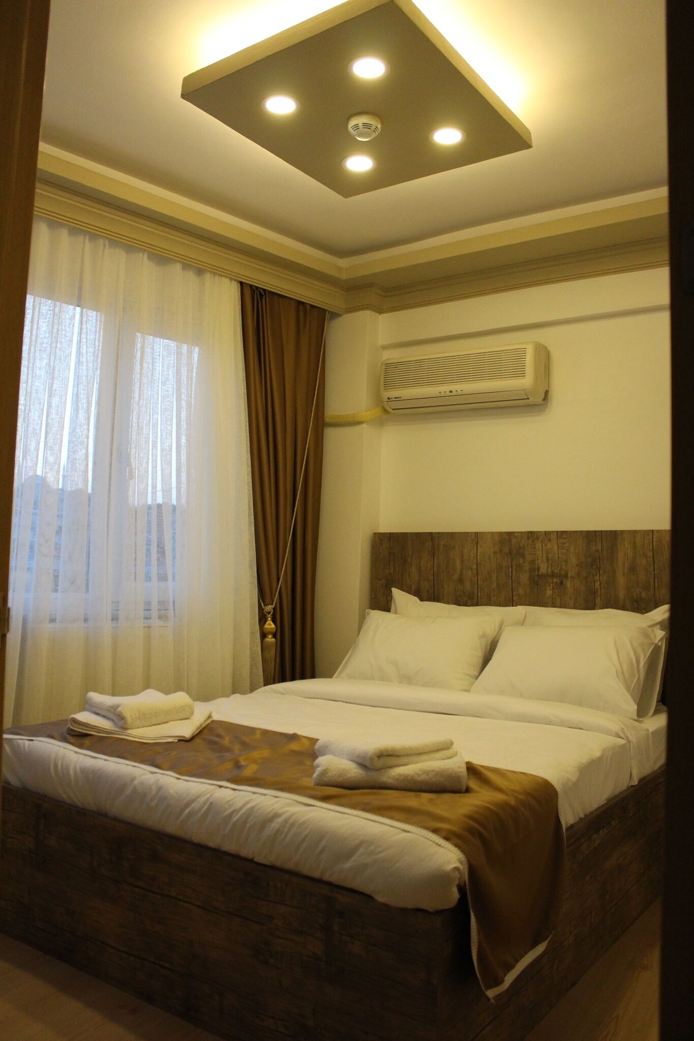 A hotel room with a bed, curtains, an air conditioning unit, and a ceiling light fixture.