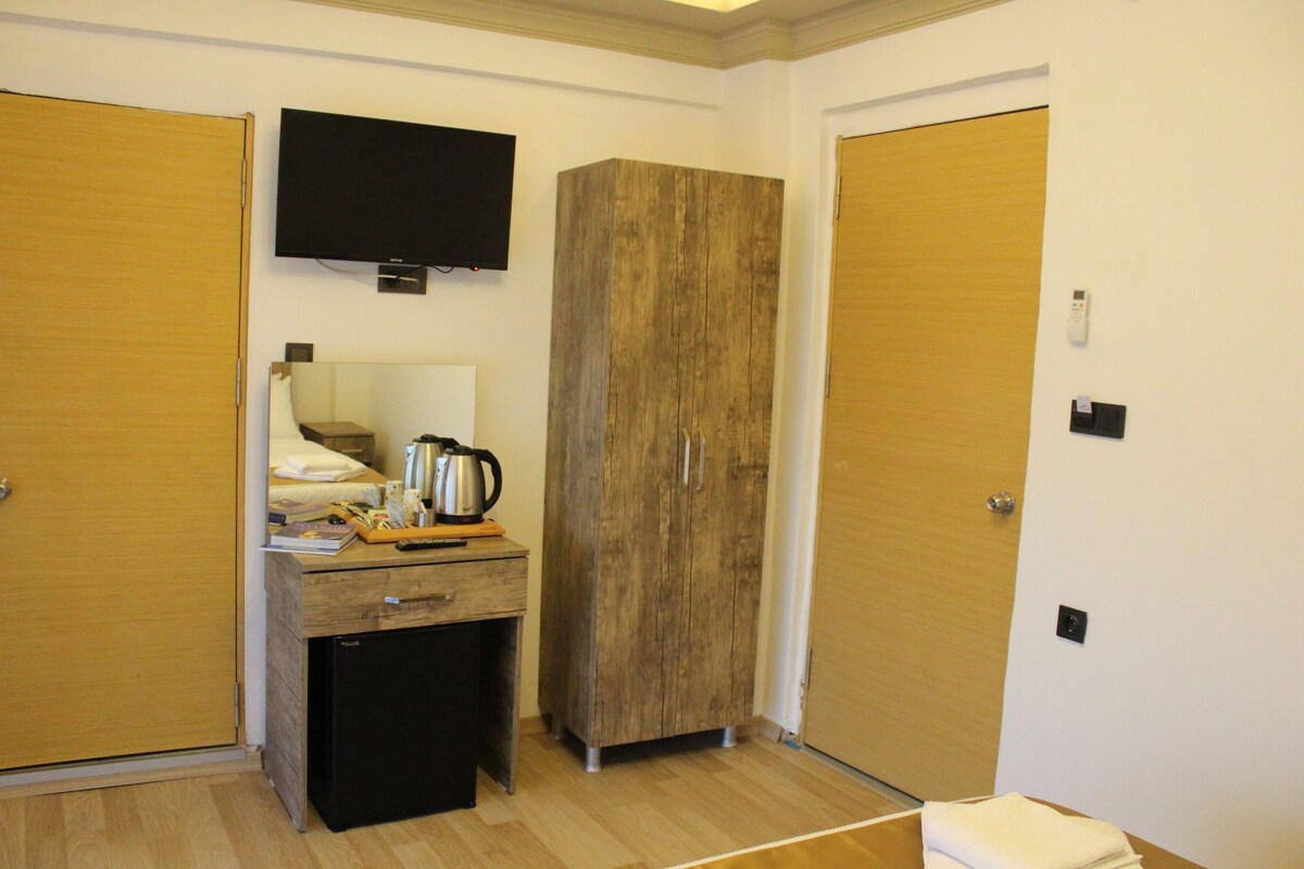 Habitació d'hotel amb un armari de fusta, un petit escriptori amb un bullidor, un televisor muntat sobre l'armari, i una porta de fusta.