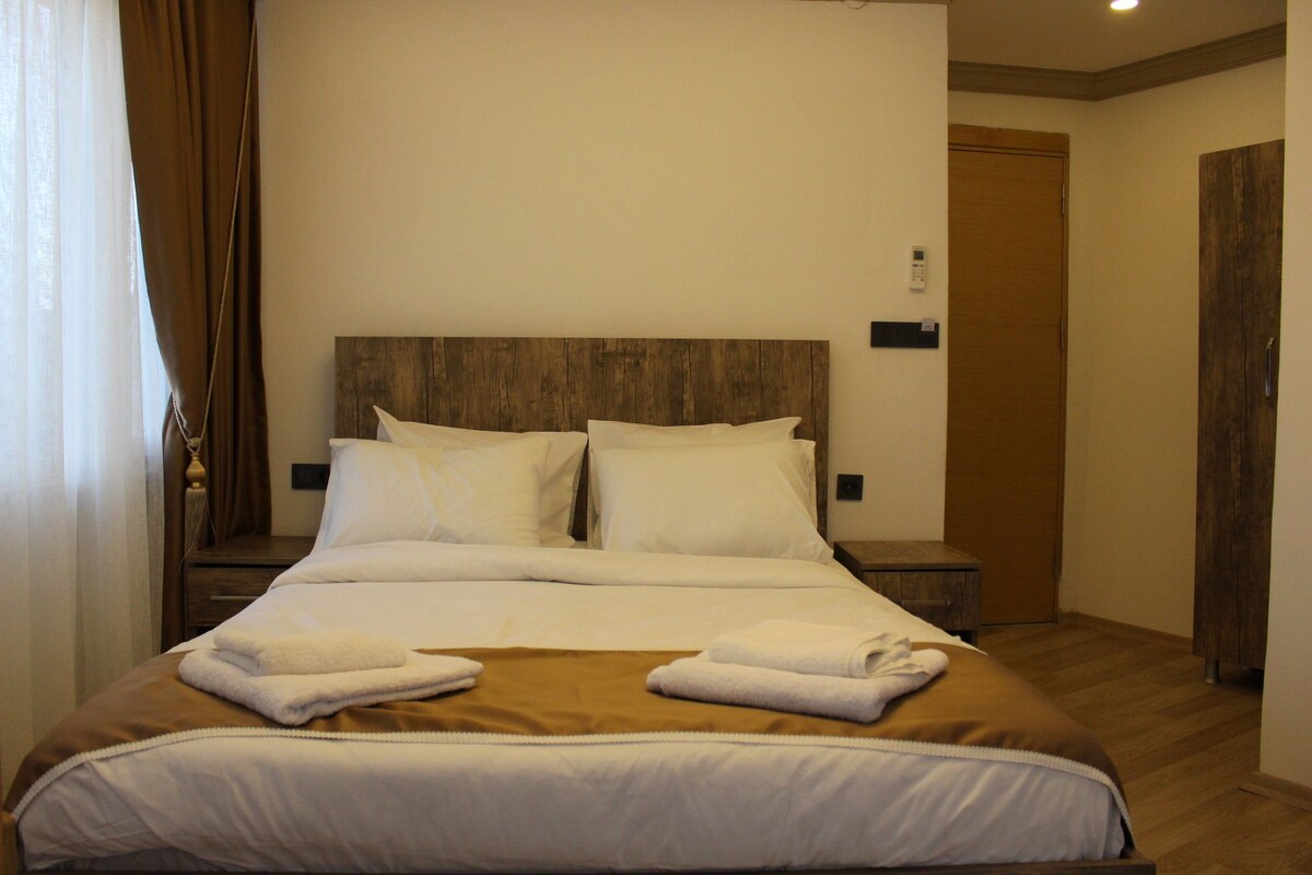 Habitació d'hotel amb un llit gran, dues taules de nit, un panell de fusta al capçal i una porta.
