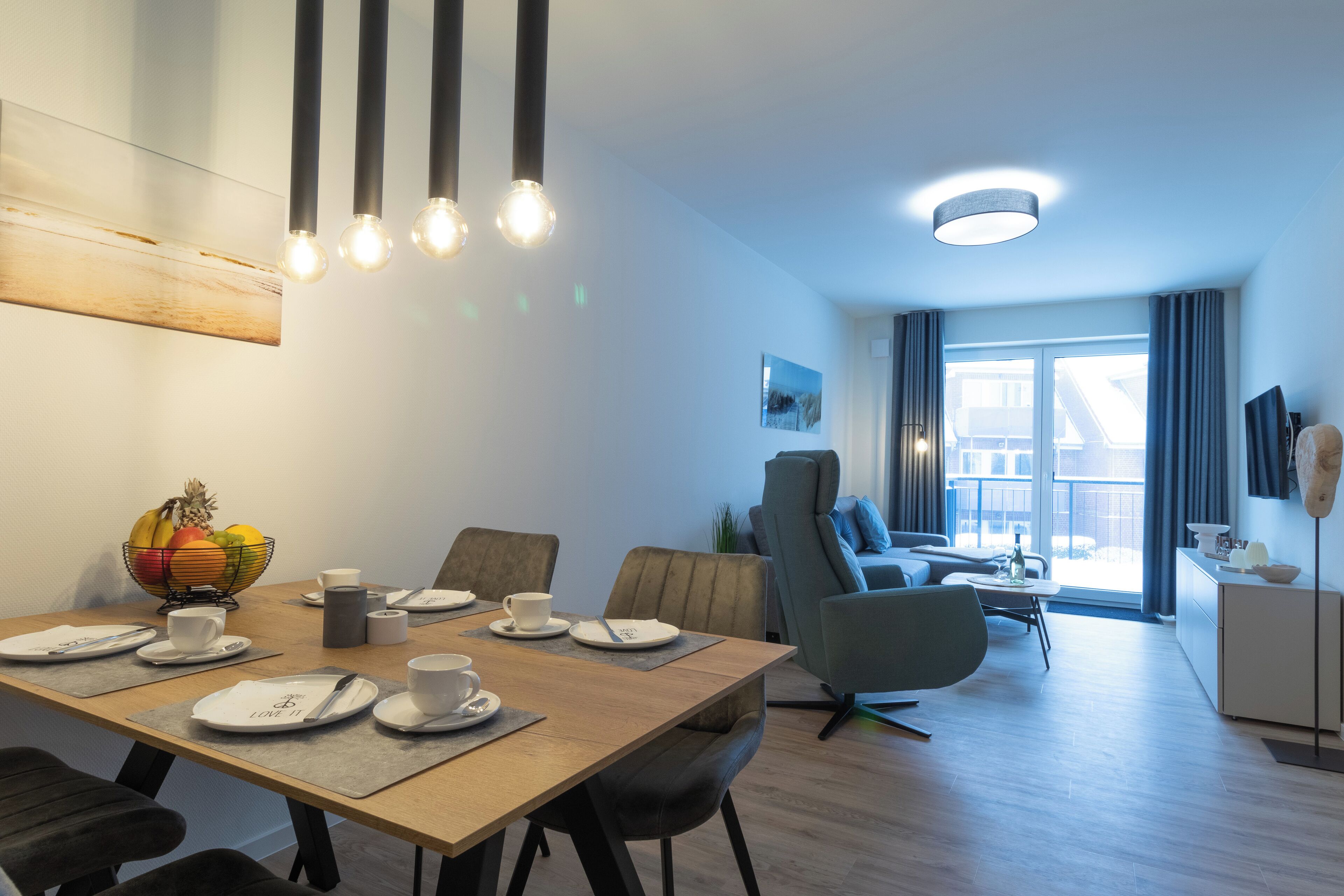 Ein moderner Essbereich mit einem Holztisch, gedeckt für vier Personen, einer Obstschale und Blick auf das Wohnzimmer mit Sofa und Balkon.