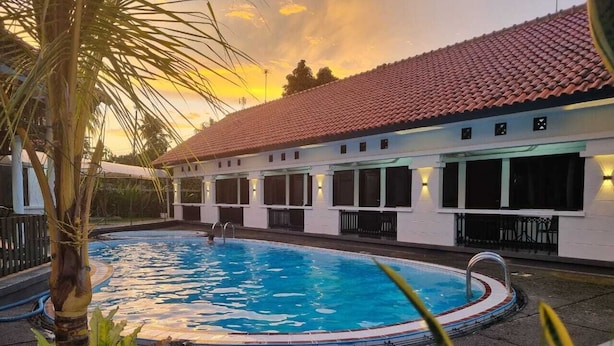 A swimming pool with a tiled roof and white walls, surrounded by plants and a palm tree. - Hotel pondok putri (Cijulang)