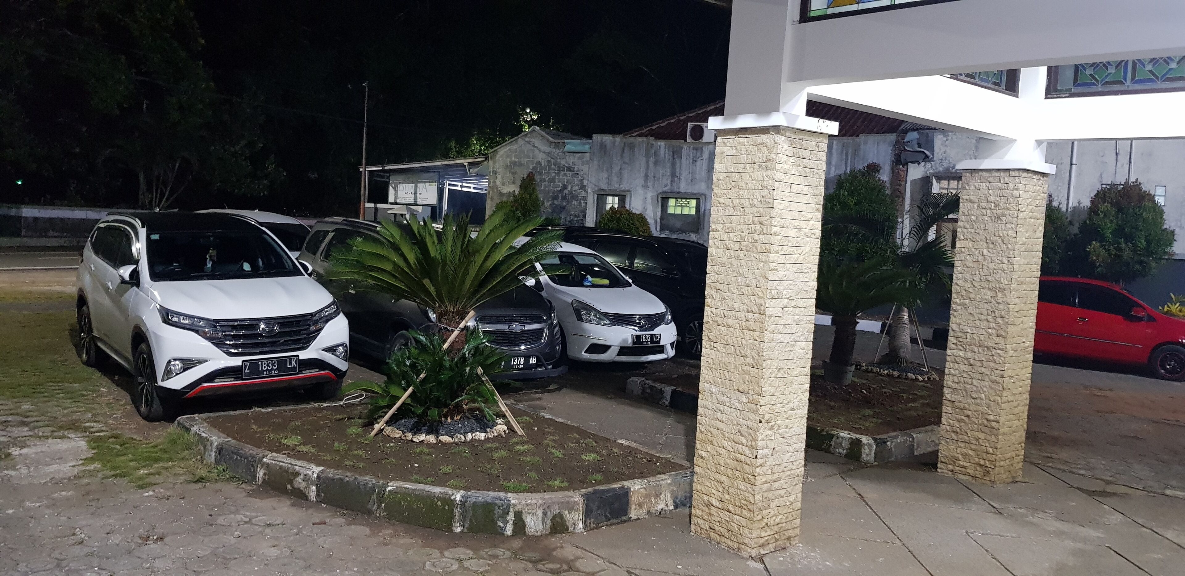 A parking area with several cars, a small garden with a palm tree, and a building with a stone facade.