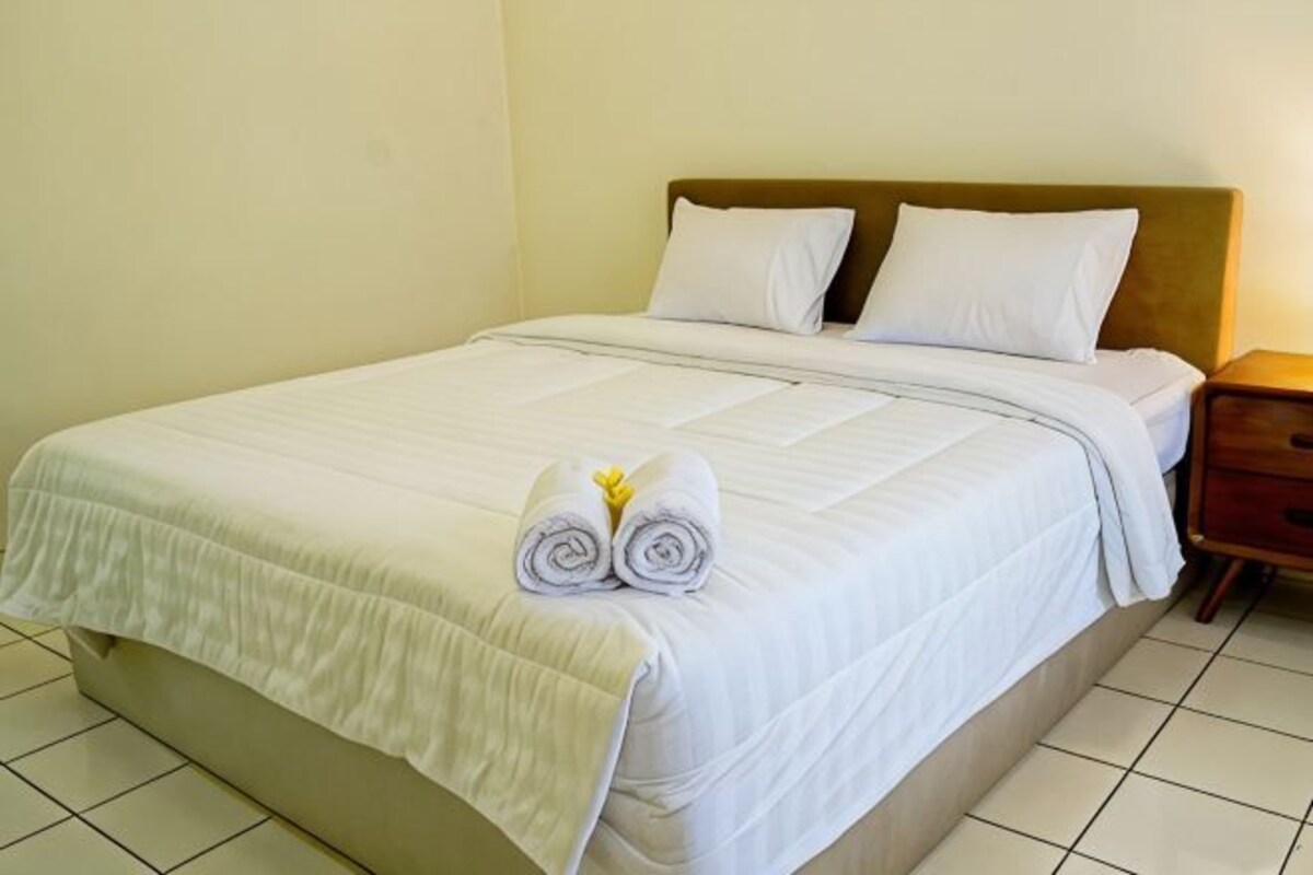 Una cama bien hecha con sábanas blancas y dos almohadas, un cabecero de madera y una mesita de noche con una lámpara.