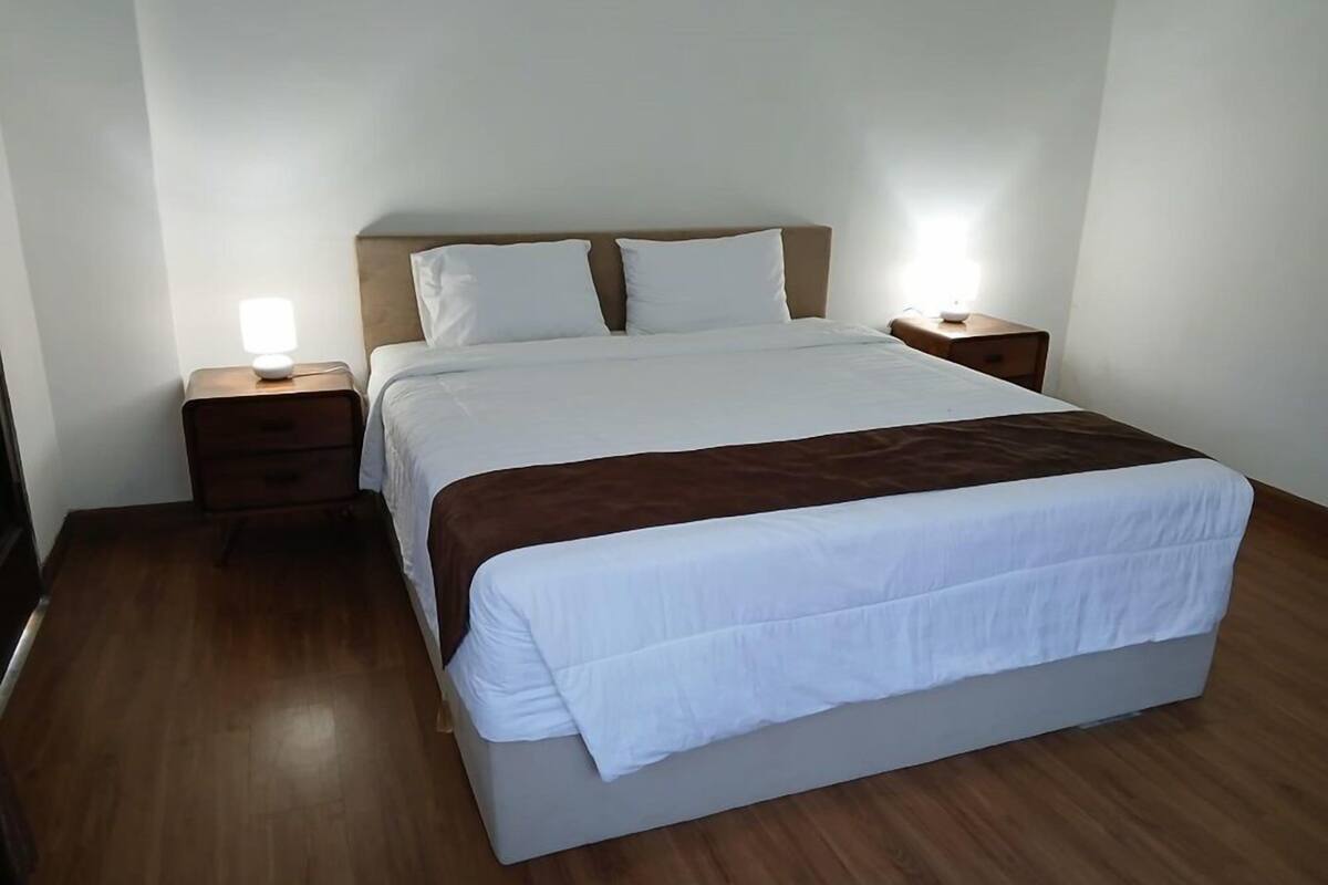 Ein ordentlich bezogenes Bett mit weißen Leinen und einer braunen Tagesdecke, zwei Nachttische mit Lampen und ein Holzboden.