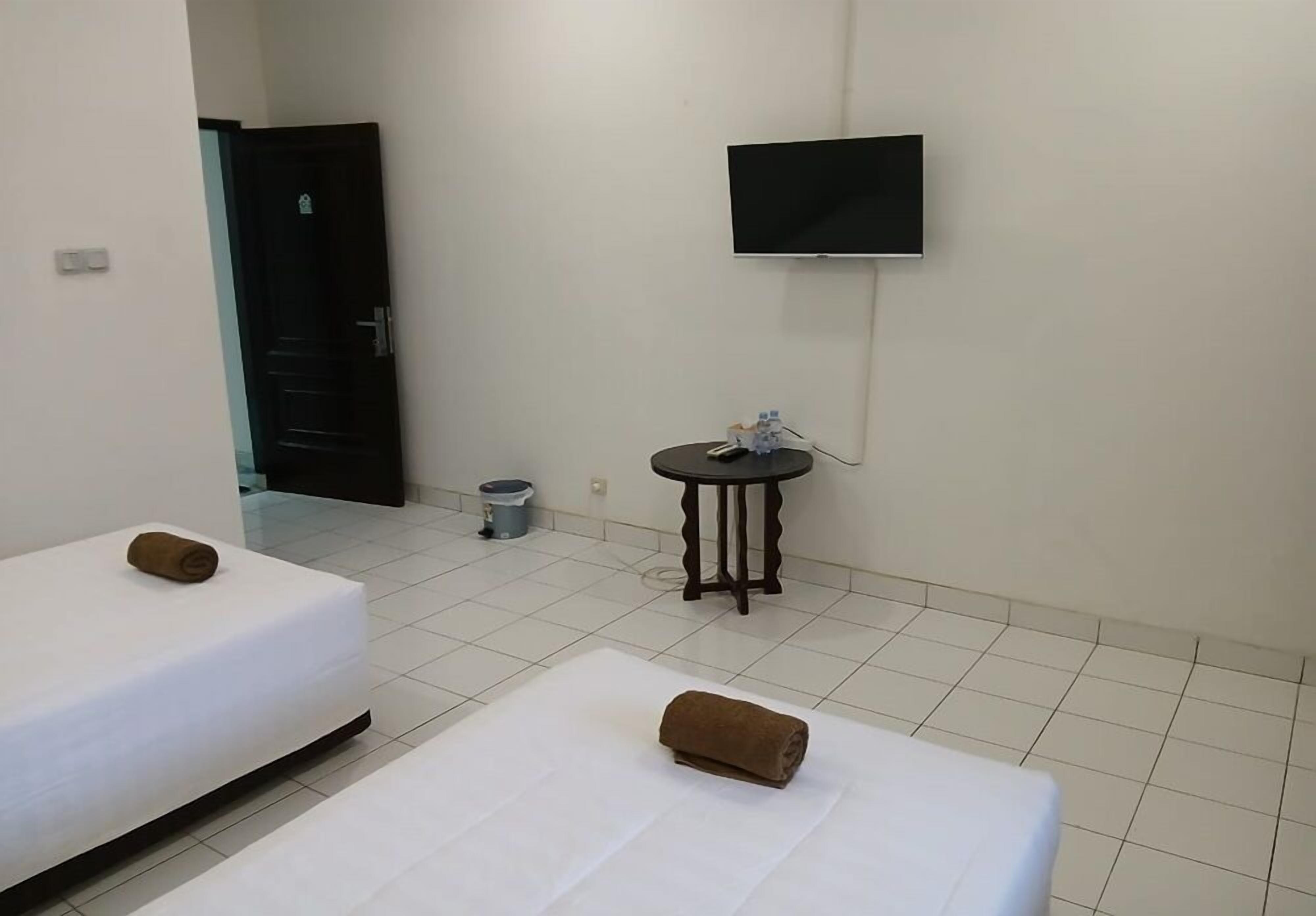 Habitación de hotel con dos camas, un televisor montado en la pared, una mesita con una botella y un cesto de basura.