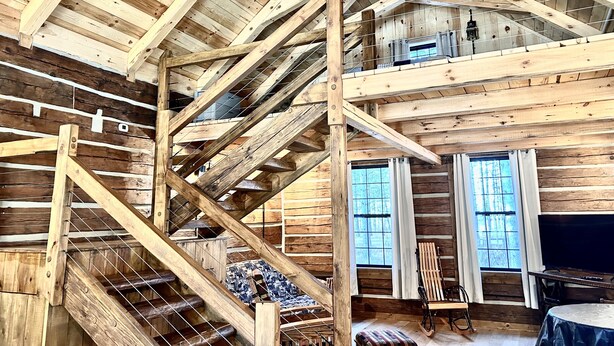 A spacious log cabin interior with a high ceiling, a wooden staircase, a loft area, and large windows.