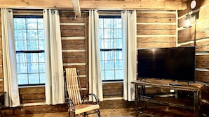A log cabin interior with a rocking chair, a television on a wooden stand, and large windows with curtains.