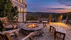 An outdoor patio with a stone fire pit, wooden chairs, and a view of a forested area. - Ski-In Ski-Out•Hot Tub•King Bed•Sauna•Deck (Snowshoe)