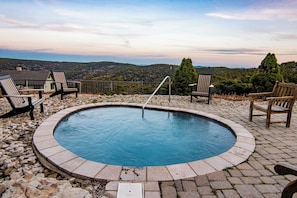 Uma piscina circular com uma escada de metal, rodeada por ladrilhos de pedra e cadeiras de madeira.