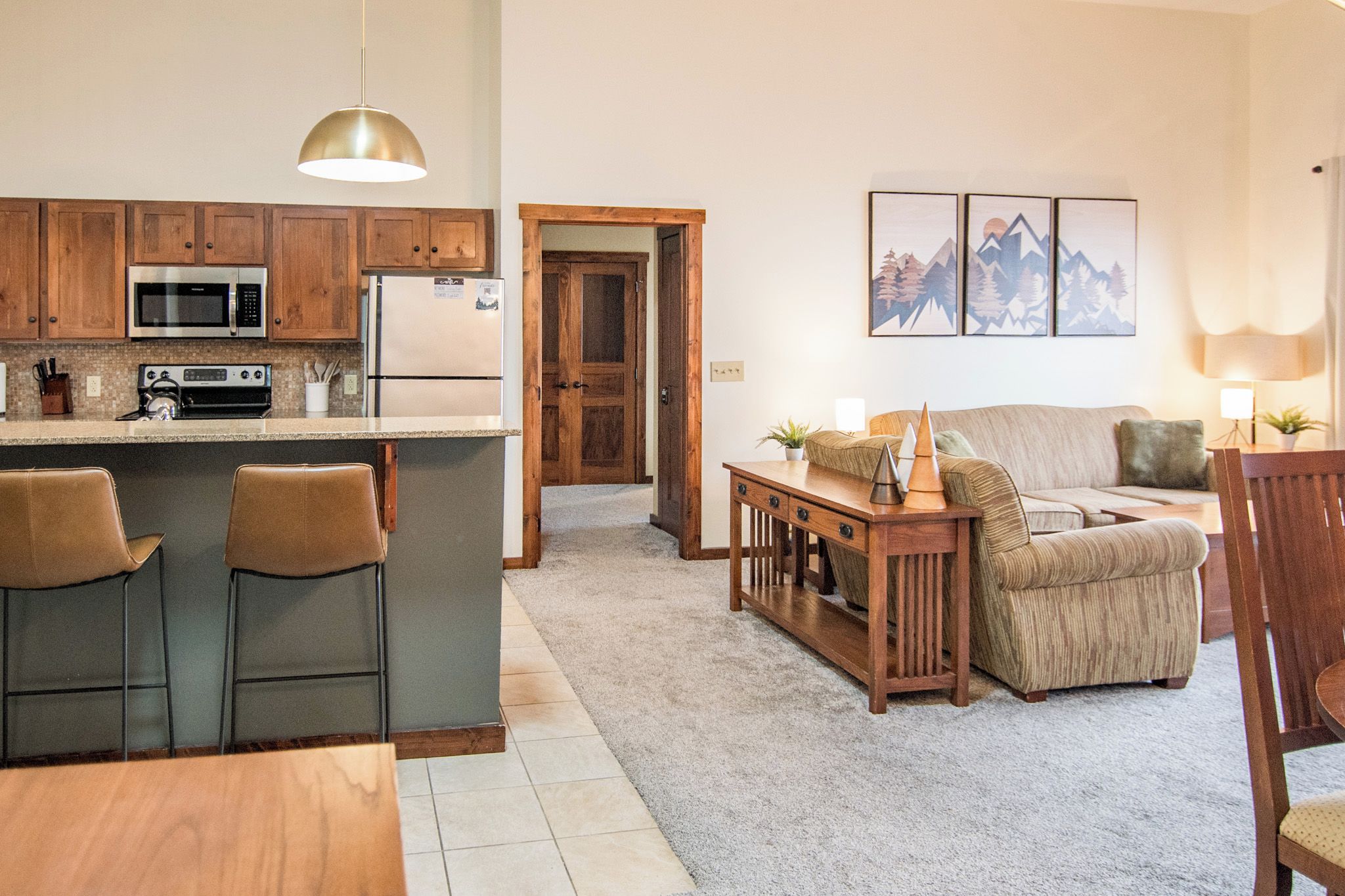 Een keuken met een magnetron, fornuis en koelkast, een eetgedeelte met een tafel en stoelen, en een woonkamer met een bank en een lamp.