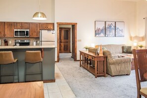 Een keuken met een magnetron, fornuis en koelkast, een eetgedeelte met een tafel en stoelen, en een woonkamer met een bank en een lamp.