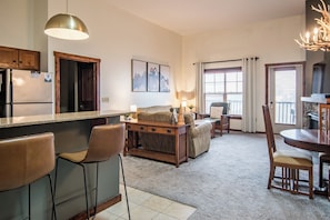 Cozinha com geladeira, armários de madeira, área de refeições com mesa e cadeiras, e sala de estar com sofá e abajur.
