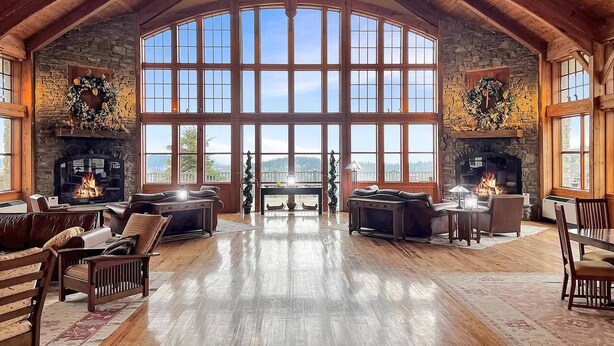 A spacious living room with a high ceiling and large windows, featuring a stone fireplace and a view of the outdoors. - Ski-In Ski-Out•Hot Tub•King Bed•Sauna•Deck (Snowshoe)