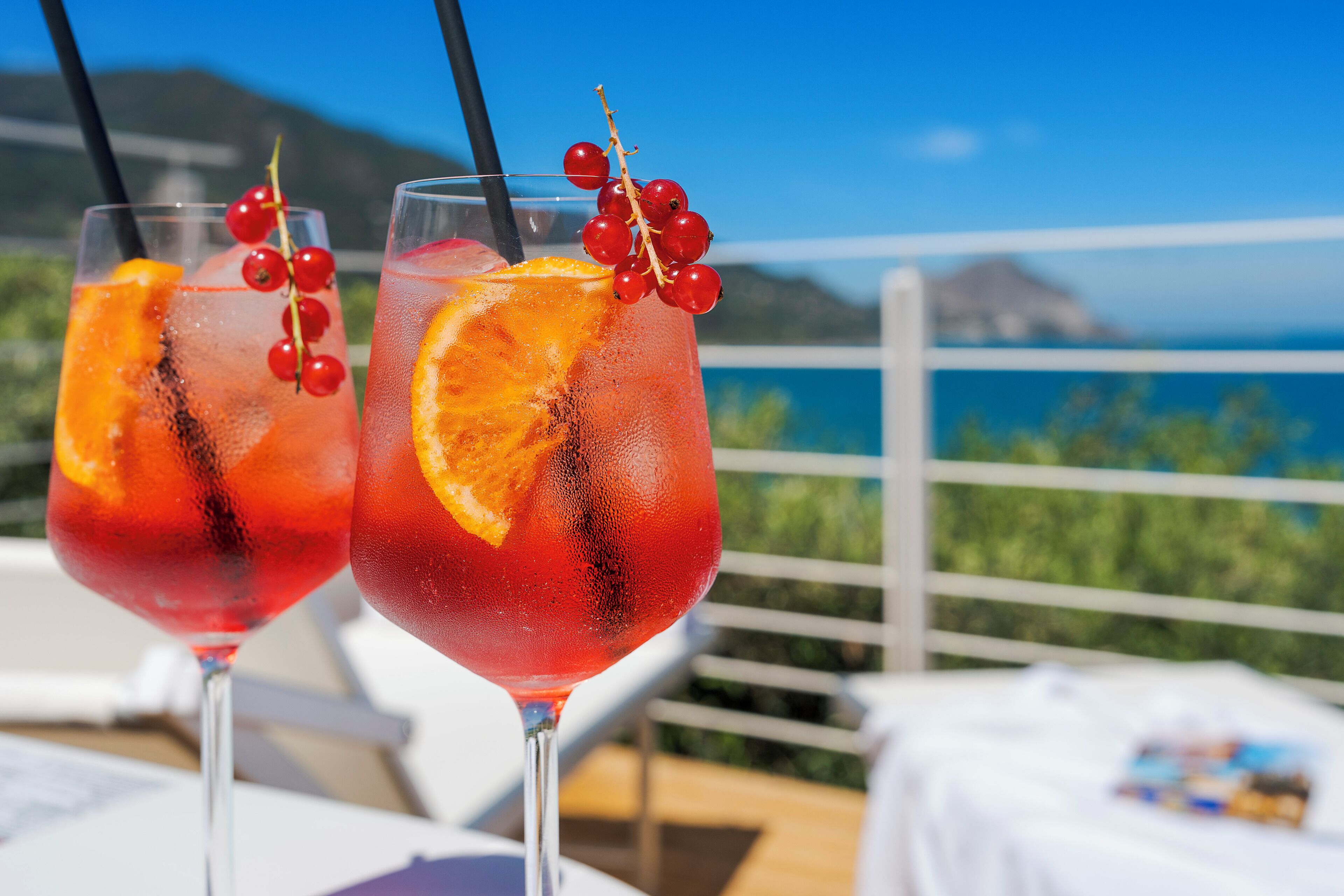 Two glasses of red beverage with orange slices and red berries, set on a balcony with a scenic view.