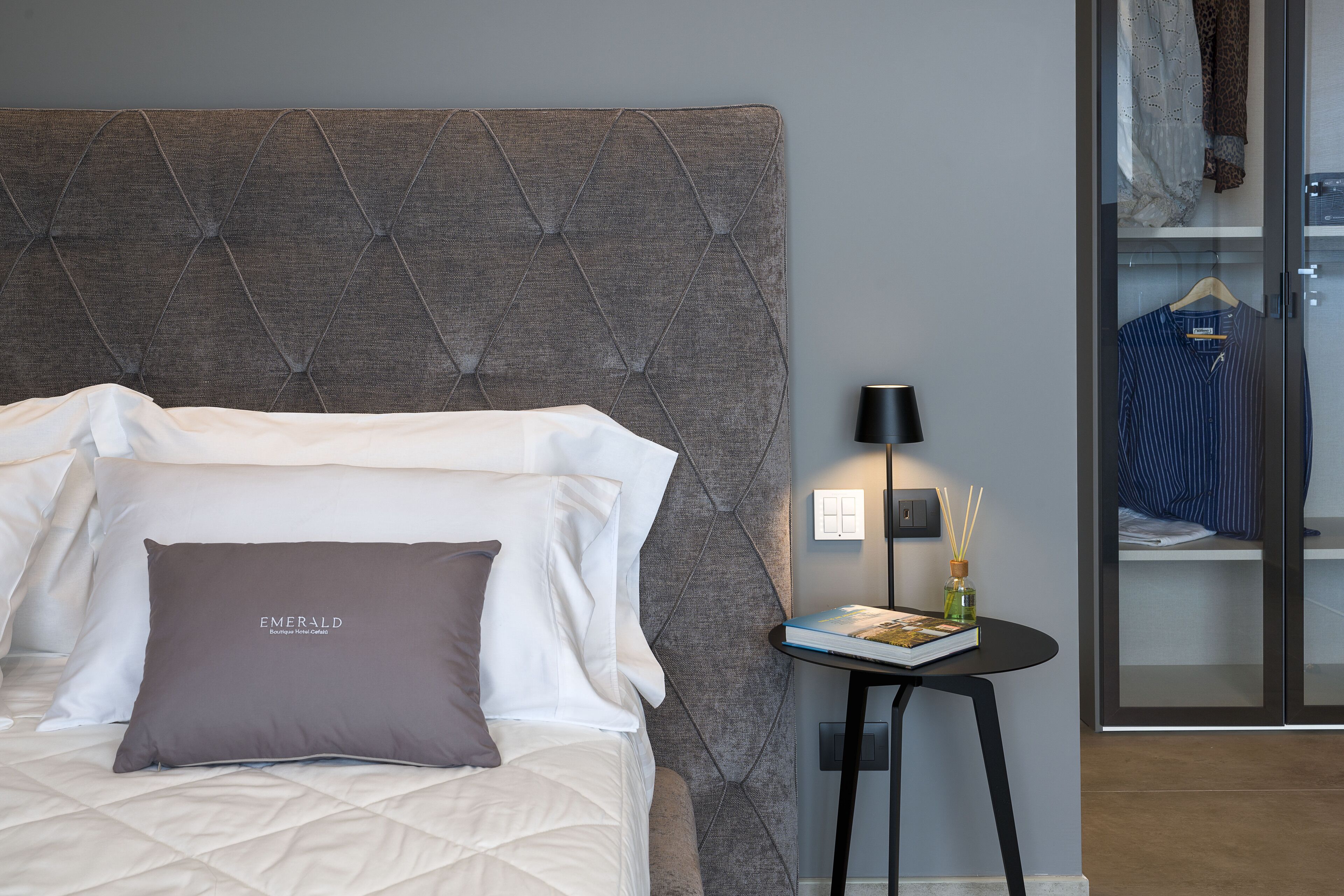 A neatly made bed with white and brown pillows, a grey upholstered headboard, a small table with a book and a bottle of water, and a clothing rack with hanging garments.