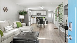 A modern living room with a grey sofa, black ottoman, and a dining area with a table and chairs. A kitchen with white cabinets and a microwave is visible in the background. - Hollywood of the south 3bed 2bath, Short walk to the square (Covington)