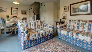 Sala de estar com sofás estampados com flores, uma mesa de jantar com cadeiras, uma escada e quadros nas paredes.