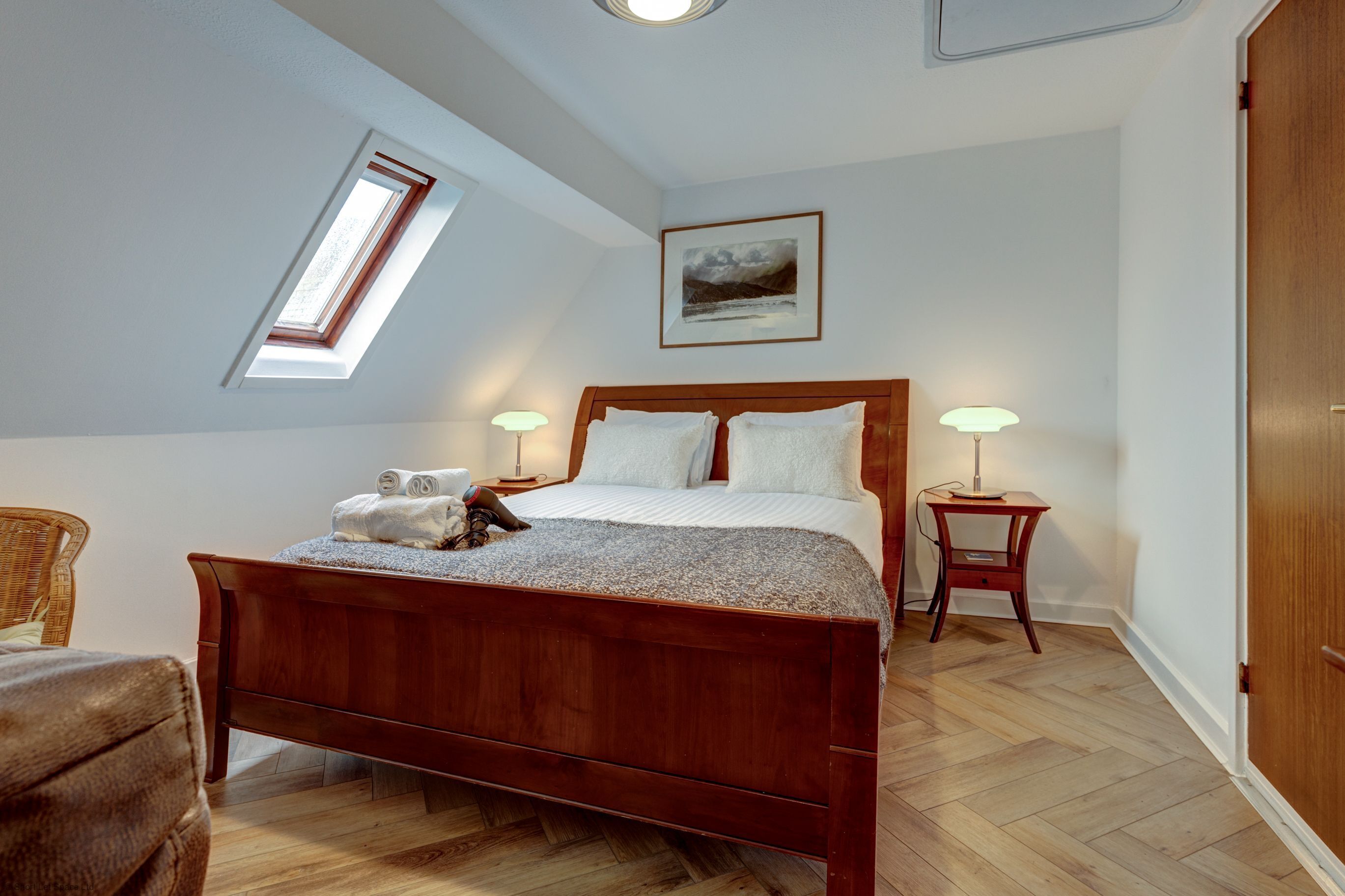 A bedroom with a wooden bed, a skylight, a bedside table with a lamp, and a framed picture on the wall.